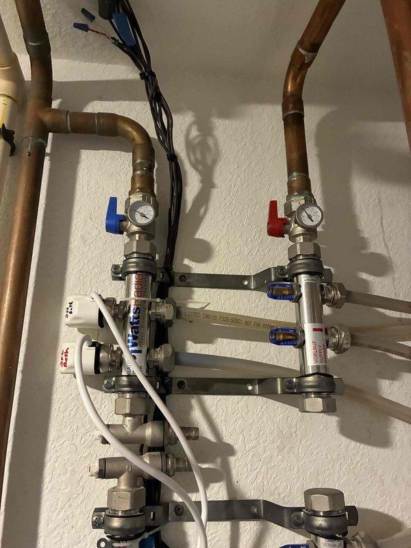 Responded to service call for in-floor radiant heating system with suspected zone control issue where bathroom zone was not calling for heat while another zone was receiving excessive heat flow. Inspected manifold assembly with pressure gauges, zone valves, and supply/return piping to diagnose zone switching problem. Documented hydronic heating manifold configuration including blue and red handled ball valves, pressure gauges, and white PEX distribution lines connected to the zone control system for troubleshooting and future reference.