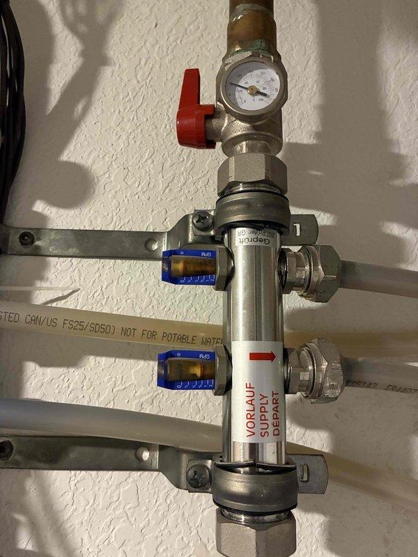 Responded to service call for in-floor radiant heating system with suspected zone control issue where bathroom zone was not calling for heat while another zone was receiving excessive heat flow. Inspected manifold assembly with pressure gauges, zone valves, and supply/return piping to diagnose zone switching problem. Documented hydronic heating manifold configuration including blue and red handled ball valves, pressure gauges, and white PEX distribution lines connected to the zone control system for troubleshooting and future reference.