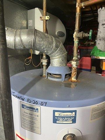 Conducted comprehensive assessment of 20+ year old Kenmore water heater requiring replacement. Unit shows significant age deterioration with installation date noted as "10-20-07" and is located in confined utility space with existing galvanized metal ductwork and copper supply lines. Provided customer with detailed replacement options and cost estimate for complete system upgrade.