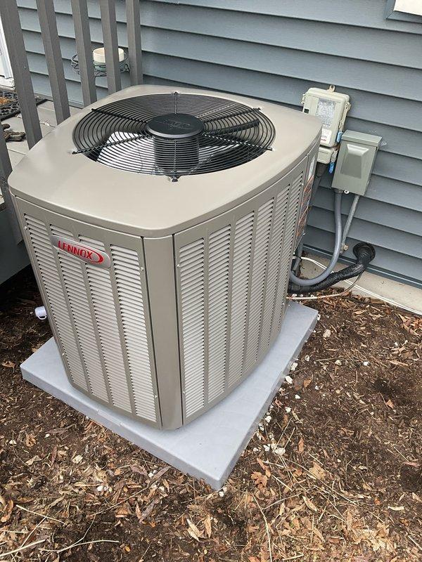 Completed installation of Lennox EL15CK1-036 air conditioning condenser unit on existing concrete pad against exterior wall with light blue vinyl siding. Unit positioned with proper clearance for airflow and service access, connected to electrical disconnect box mounted on exterior wall via conduit. AC system paired with furnace and coil previously installed in October 2025 to complete staged HVAC installation. Unit tested and operational, with 27A03 module noted for future installation to finalize system setup.
