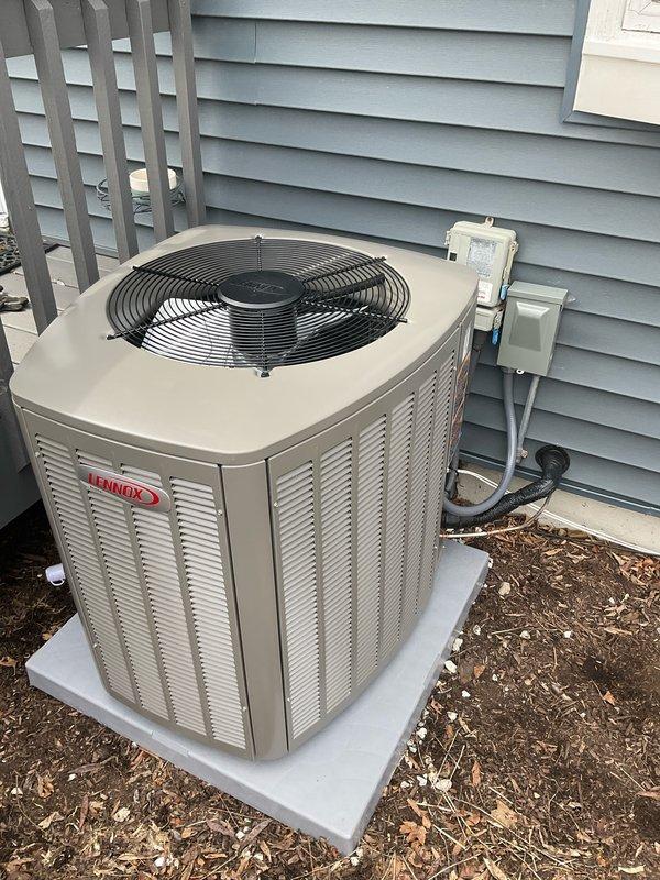 Completed installation of Lennox EL15CK1-036 air conditioning condenser unit on existing concrete pad against exterior wall with light blue vinyl siding. Unit positioned with proper clearance for airflow and service access, connected to electrical disconnect box mounted on exterior wall via conduit. AC system paired with furnace and coil previously installed in October 2025 to complete staged HVAC installation. Unit tested and operational, with 27A03 module noted for future installation to finalize system setup.