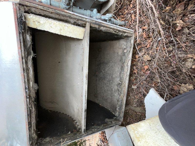 Performed site assessment of rooftop commercial air conditioning system to provide replacement estimate for condenser and evaporator coil. Accessed rooftop mechanical equipment through concrete-lined compartment with weathered access panel showing signs of age and corrosion. Inspected ductwork plenum and air handling components, noting fibrous insulation material with visible wear patterns and multiple mounting fasteners. Documented existing system configuration including galvanized metal panels and insulation conditions within the compartment. Photographed equipment access points and internal ductwork components to support estimate preparation for complete condenser and coil replacement on commercial HVAC system.