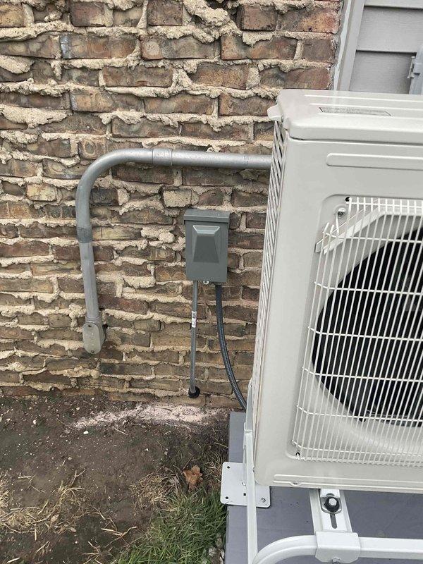 Completed new installation of 9,000 BTU Mitsubishi Hyper Heat mini-split system with outdoor condenser unit mounted on white metal stand against exterior brick and siding. Installed 15-foot refrigerant line set and 15-foot electrical conduit run, connecting outdoor unit to indoor air handler with gray PVC conduit housing and electrical disconnect box mounted to exterior wall. Total installation time was 12 hours, resulting in fully operational heating and cooling system.