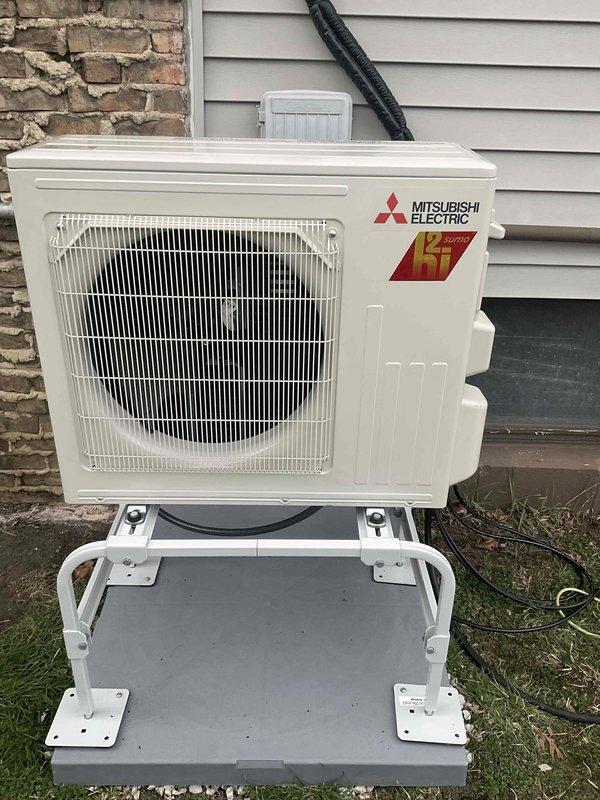 Completed new installation of 9,000 BTU Mitsubishi Hyper Heat mini-split system with outdoor condenser unit mounted on white metal stand against exterior brick and siding. Installed 15-foot refrigerant line set and 15-foot electrical conduit run, connecting outdoor unit to indoor air handler with gray PVC conduit housing and electrical disconnect box mounted to exterior wall. Total installation time was 12 hours, resulting in fully operational heating and cooling system.