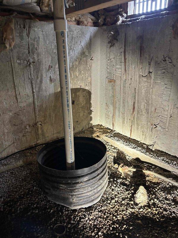 Replaced non-functional sump pump system in residential crawl space. Removed existing failed pump and basin from deteriorated below-grade area showing significant water damage and moisture infiltration on concrete walls. Installed new sump pump with black ribbed basin positioned on gravel floor and connected white PVC discharge line extending vertically through crawl space. Verified discharge pipe properly routes water away from foundation exterior, with horizontal PVC outlet positioned near ground level on building perimeter. System tested and confirmed operational for effective groundwater management and foundation protection.