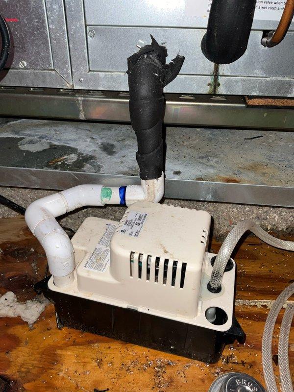Responded to service call for water leak from attic-mounted air conditioning system causing water intrusion into basement. Upon inspection, found condensate pump experiencing drainage issues with visible water accumulation on wooden platform beneath unit. Identified deteriorated insulation on condensate piping and evidence of ongoing moisture problems around pump assembly. Documented existing conditions including damaged pipe insulation, water staining on plywood surface, and condensate pump installation requiring attention to resolve leak and prevent further water damage.