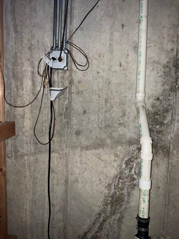 Responded to service call for sump pump issues in residential basement. Upon inspection, assessed sump pump system including discharge piping, electrical connections, and pit condition. Evaluated white PVC discharge line with wall-mounted routing and black coupling adapter at floor penetration point. Inspected electrical junction box and associated wiring serving the pump system. Documented basement moisture conditions including wall staining and efflorescence on concrete surfaces, which indicated previous water intrusion. Photographed existing sump pit configuration and discharge infrastructure for service records.