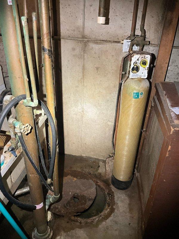 Responded to service call for aging water heater exhibiting active leak and sump pump replacement request. Upon arrival, inspected existing water heater in basement utility area and confirmed deterioration with visible moisture damage. Assessed surrounding plumbing infrastructure including copper supply lines and existing water treatment system. Evaluated sump pump pit location showing evidence of previous water accumulation and floor staining around drainage area. Documented equipment conditions and basement mechanical room layout including overhead PVC discharge piping and flexible connector installations. Provided recommendations for water heater replacement and sump pump upgrade to prevent future water damage and ensure proper basement drainage functionality.