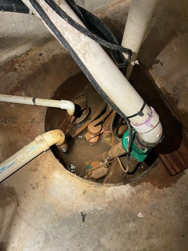 Responded to service call for malfunctioning sump pump with incomplete water removal and excessive noise during operation. Upon arrival, performed diagnostic inspection of sump pump system located in basement utility area. Found pump basin containing elevated water level with murky brown water, indicating inadequate discharge performance. Inspected green cylindrical sump pump unit positioned in concrete basin along with associated PVC discharge piping, inlet connections, and mounting hardware. Evaluated pump motor operation, float switch mechanism, and check valve function. Assessed vertical discharge line, pipe fittings, clamps, and support straps for proper installation and potential blockages. Examined electrical connections at wall-mounted outlet box to verify power supply integrity. Documented system configuration including white PVC piping network, basin access point, and equipment positioning. Identified operational deficiencies contributing to reported symptoms and provided rec
