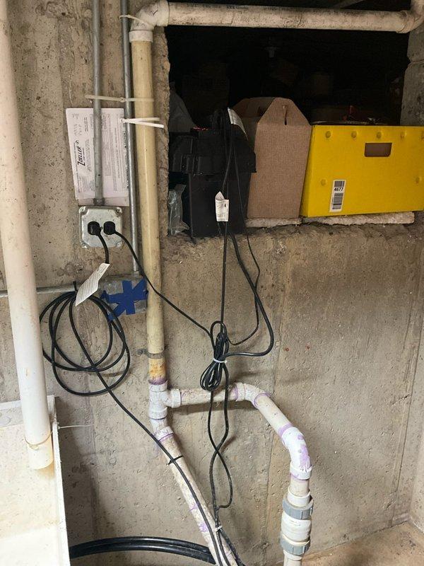 Responded to service call for malfunctioning sump pump with incomplete water removal and excessive noise during operation. Upon arrival, performed diagnostic inspection of sump pump system located in basement utility area. Found pump basin containing elevated water level with murky brown water, indicating inadequate discharge performance. Inspected green cylindrical sump pump unit positioned in concrete basin along with associated PVC discharge piping, inlet connections, and mounting hardware. Evaluated pump motor operation, float switch mechanism, and check valve function. Assessed vertical discharge line, pipe fittings, clamps, and support straps for proper installation and potential blockages. Examined electrical connections at wall-mounted outlet box to verify power supply integrity. Documented system configuration including white PVC piping network, basin access point, and equipment positioning. Identified operational deficiencies contributing to reported symptoms and provided rec