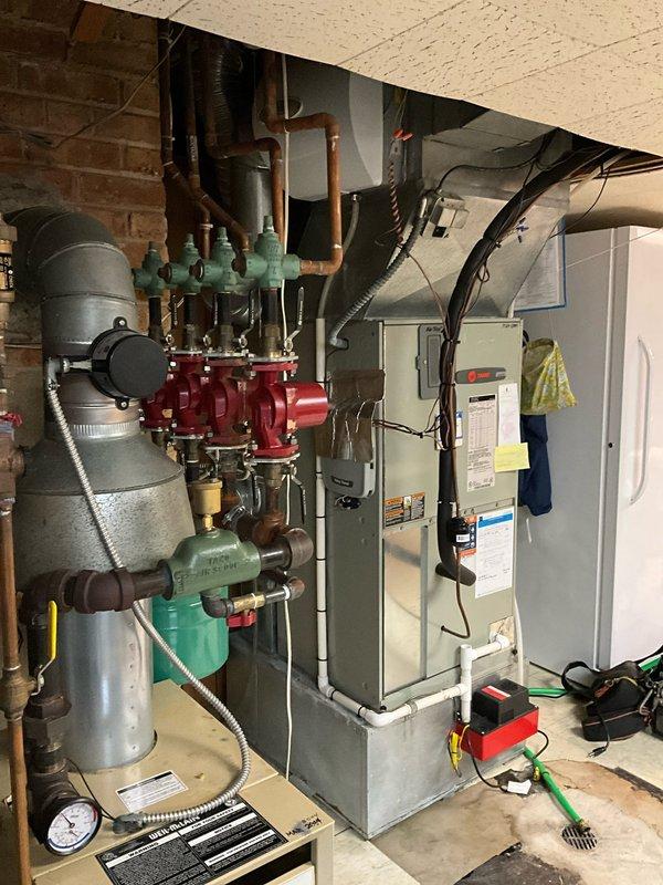 Responded to after-hours emergency call for water leaking from AC unit with malfunctioning condensate pump and non-responsive thermostat. Upon arrival, inspected wall-mounted digital thermostat displaying blank screen and confirmed complete loss of power to control unit, preventing system shutdown. Proceeded to basement mechanical room and evaluated HVAC system including furnace, multi-zone manifold assembly with green valve controls, and floor-mounted condensate pump with pressure gauge. Identified condensate pump failure as primary cause of water leakage and thermostat malfunction related to electrical issue. Documented equipment conditions including copper and PEX piping connections, red manifold distribution system, expansion tank, and associated electrical conduits for further diagnostic evaluation and repair recommendations.
