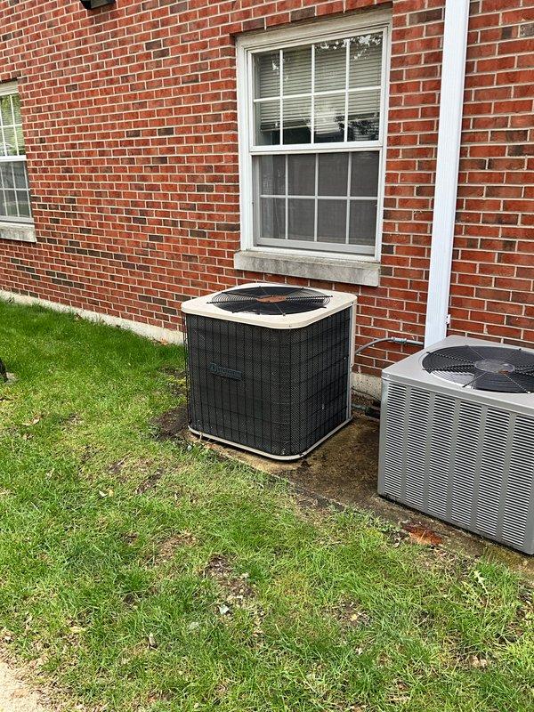 Conducted comprehensive full system HVAC estimate at residential property with brick exterior. Evaluated existing dual outdoor condenser units positioned on ground-level installation adjacent to building foundation, with visible signs of aging including rust staining and corrosion on mounting surfaces and connection points. Assessed condensate drainage system consisting of white PVC piping in U-shaped configuration and metallic flex connectors showing deterioration. Documented current equipment condition, noting surface wear on concrete pad, discoloration around pipe connections, and overall system age to provide accurate replacement recommendations for complete heating and cooling system upgrade.
