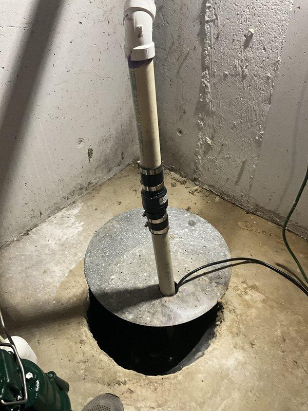 Removed and disposed of existing sump pump and check valve assembly from basement sump pit. The original pump was extracted from the circular concrete pit, which featured a split concrete cover to accommodate the discharge piping. Disconnected the white PVC discharge pipe with black rubber check valve components and removed the wall-mounted bracket securing the vertical pipe run. Verified proper drainage routing from the basement through exterior discharge lines, which terminate in the mulched area alongside the building foundation with black corrugated extension pipes directing water away from the structure.