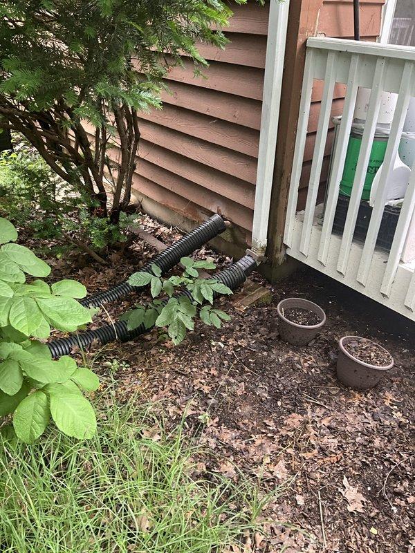Removed and disposed of existing sump pump and check valve assembly from basement sump pit. The original pump was extracted from the circular concrete pit, which featured a split concrete cover to accommodate the discharge piping. Disconnected the white PVC discharge pipe with black rubber check valve components and removed the wall-mounted bracket securing the vertical pipe run. Verified proper drainage routing from the basement through exterior discharge lines, which terminate in the mulched area alongside the building foundation with black corrugated extension pipes directing water away from the structure.