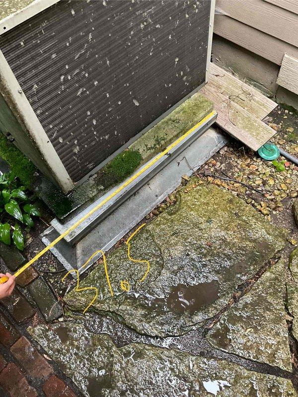 Conducted site assessment for mini split system installation estimate. Evaluated exterior location near existing HVAC equipment, noting adequate space for outdoor condenser unit placement on flagstone paver area adjacent to building with brown horizontal siding. Documented current drainage conditions including standing water in existing drip pan and moss growth on pavers, which will require consideration for proper condensate management. Performed interior measurements to determine optimal indoor air handler placement and assess wall penetration requirements for refrigerant line set routing. Measured available vertical clearance and documented wall construction to plan line hide installation path. Captured photographic documentation of proposed installation locations both exterior and interior to develop accurate equipment sizing recommendations and installation scope.