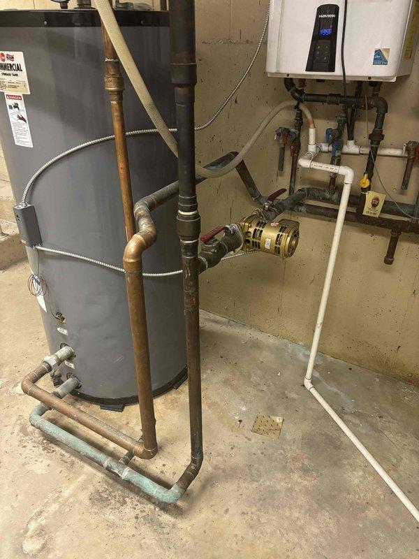 Returned to property to assess and provide estimate for piping repair on water heater manifold system. Upon inspection, evaluated the dual water heating setup consisting of a traditional tank-style water heater and wall-mounted tankless unit with interconnected copper and PVC piping. Examined the manifold assembly featuring multiple ball valves, including blue and yellow-handled shutoff valves, flexible braided connectors, and various T-joints and elbow fittings. Documented weathered metal piping showing signs of age and corrosion, as well as the configuration of supply and distribution lines. Photographed the complete plumbing infrastructure to prepare comprehensive repair quote for necessary manifold piping corrections.