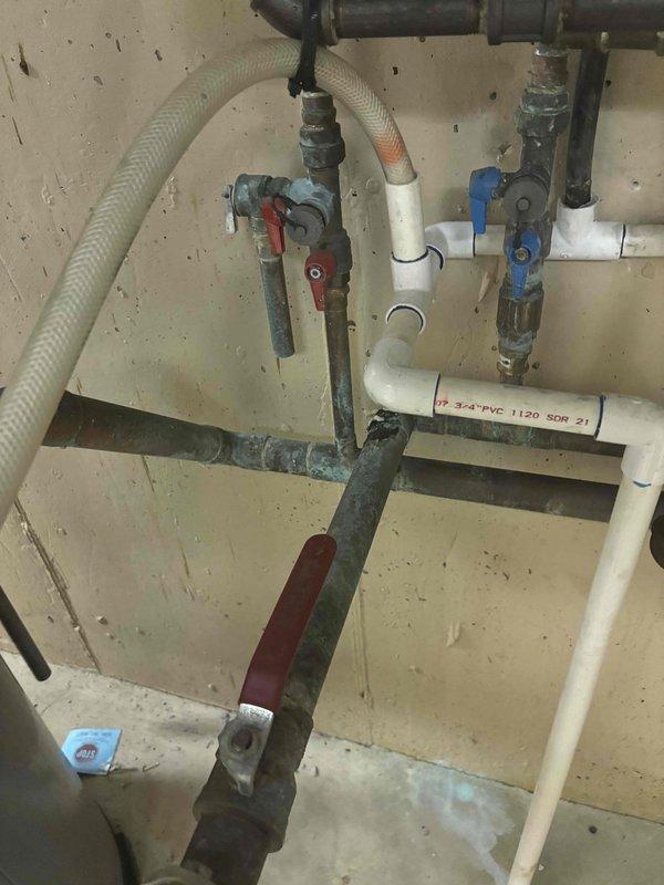 Returned to property to assess and provide estimate for piping repair on water heater manifold system. Upon inspection, evaluated the dual water heating setup consisting of a traditional tank-style water heater and wall-mounted tankless unit with interconnected copper and PVC piping. Examined the manifold assembly featuring multiple ball valves, including blue and yellow-handled shutoff valves, flexible braided connectors, and various T-joints and elbow fittings. Documented weathered metal piping showing signs of age and corrosion, as well as the configuration of supply and distribution lines. Photographed the complete plumbing infrastructure to prepare comprehensive repair quote for necessary manifold piping corrections.