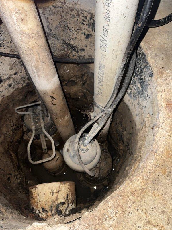 # Plumbing Service - Sump Pump

Conducted comprehensive inspection of residential sump pump system and related plumbing infrastructure. Identified active leak in waste line requiring immediate plumbing intervention. Examined sump pump installation located in concrete-lined pit, noting standing water with visible sediment and debris accumulation. Discovered battery backup system experiencing leakage with active error code displayed on control panel, indicating system malfunction. Assessed overall condition of pump mechanism, float switch assembly, and associated PVC discharge piping showing signs of age and mineral deposits. Documented heavy staining on concrete pit walls consistent with long-term water exposure. Recommended professional plumber evaluate and repair waste line leak. Advised sump pump inspection and potential replacement due to battery failure and error code indication to prevent flooding risk. Photographed bathroom area and sump pump installation for service documentatio
