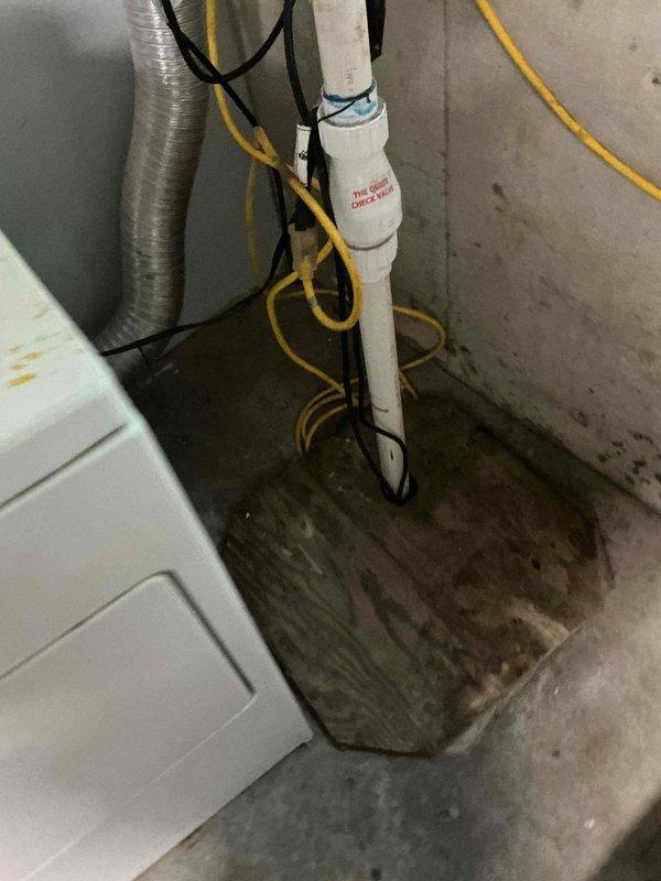 Completed comprehensive sump pump inspection in basement utility area as requested during morning hours. System evaluated including check valve operation, discharge piping, electrical connections, and sump pit condition. Floor area showed evidence of previous moisture accumulation near corner, indicating potential drainage concerns. Documented all components with photographs, verified proper installation of one-way check valve on discharge line, and assessed overall system functionality to ensure reliable operation during future water events.