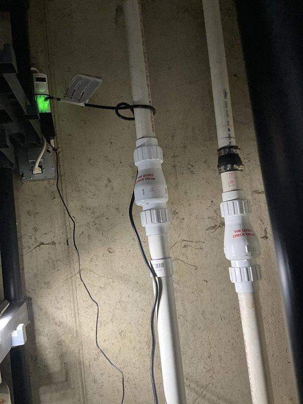 Installed battery backup sump pump system with dual pump configuration in existing sump pit. Primary and backup pumps were positioned in perforated liner with check valves installed on discharge lines to prevent backflow. White PVC discharge piping was secured to ceiling joists with proper support brackets and routed to exterior drainage point. Battery backup control unit was mounted to wall with green indicator light confirming operational status and charging function. All electrical connections were completed, system was tested for proper operation, and pit was verified for adequate water level activation.