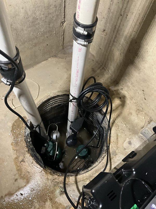 Installed battery backup sump pump system with dual pump configuration in existing sump pit. Primary and backup pumps were positioned in perforated liner with check valves installed on discharge lines to prevent backflow. White PVC discharge piping was secured to ceiling joists with proper support brackets and routed to exterior drainage point. Battery backup control unit was mounted to wall with green indicator light confirming operational status and charging function. All electrical connections were completed, system was tested for proper operation, and pit was verified for adequate water level activation.