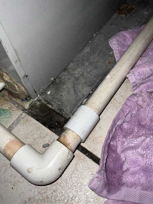 # HVAC Service Completed

Responded to service call for air conditioning unit leaking water, suspected condensate issue. Upon arrival, inspected the condensate drain system and surrounding area. Found evidence of active water leakage affecting flooring and wall surfaces in the utility space where the AC unit is located.

Examination revealed PVC condensate drain piping with visible signs of moisture and staining on pipe surfaces. Observed significant water damage to flooring tiles with gaps and cracks present, as well as moisture damage and discoloration on adjacent wall surfaces indicating ongoing or repeated water intrusion. Purple towel was present on site, apparently placed to absorb leaking water.

Traced condensate drain line routing and identified compromised drainage system allowing water to overflow or leak rather than properly discharge. The deterioration patterns on walls and flooring suggested the leak had been occurring over an extended period. 

Cleared condensate drain l