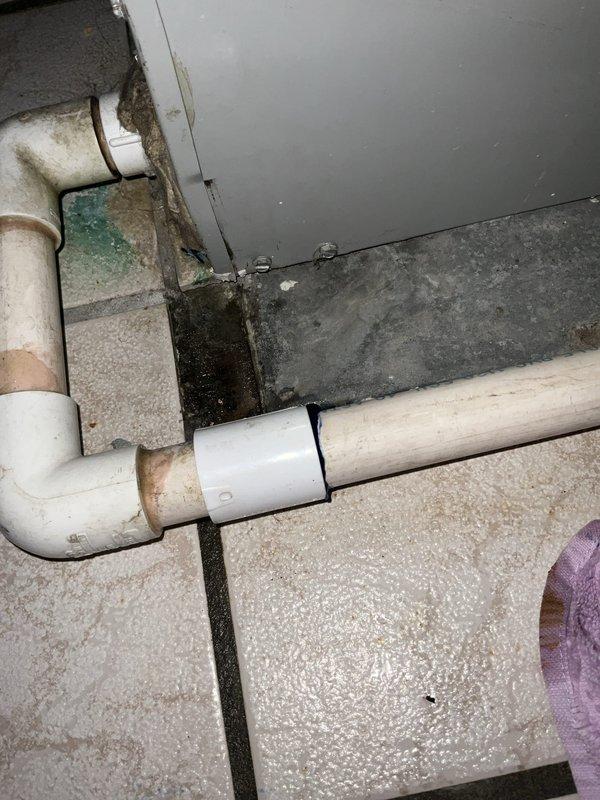 # HVAC Service Completed

Responded to service call for air conditioning unit leaking water, suspected condensate issue. Upon arrival, inspected the condensate drain system and surrounding area. Found evidence of active water leakage affecting flooring and wall surfaces in the utility space where the AC unit is located.

Examination revealed PVC condensate drain piping with visible signs of moisture and staining on pipe surfaces. Observed significant water damage to flooring tiles with gaps and cracks present, as well as moisture damage and discoloration on adjacent wall surfaces indicating ongoing or repeated water intrusion. Purple towel was present on site, apparently placed to absorb leaking water.

Traced condensate drain line routing and identified compromised drainage system allowing water to overflow or leak rather than properly discharge. The deterioration patterns on walls and flooring suggested the leak had been occurring over an extended period. 

Cleared condensate drain l