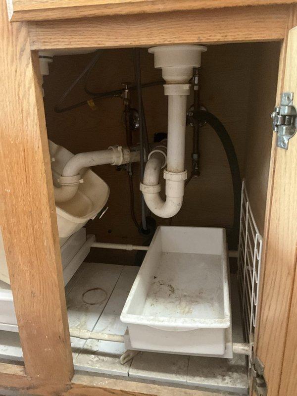 Responded to service call for water leak beneath kitchen sink at drain connection. Upon inspection, identified moisture accumulation in drip pan under cabinet, indicating active or recent leak at drain assembly. Inspected PVC P-trap and drain connections for proper seal and tightness. Addressed leak source at drain tailpiece connection point where drain exits sink basin. Tightened connections and verified proper seal at all drain assembly joints. Tested system for leaks by running water through both sink basins and monitoring drain connections for any signs of moisture. Confirmed repair successful with no active leaking observed. Cabinet interior and drip pan showed evidence of previous water exposure but no active water present after repair completion.
