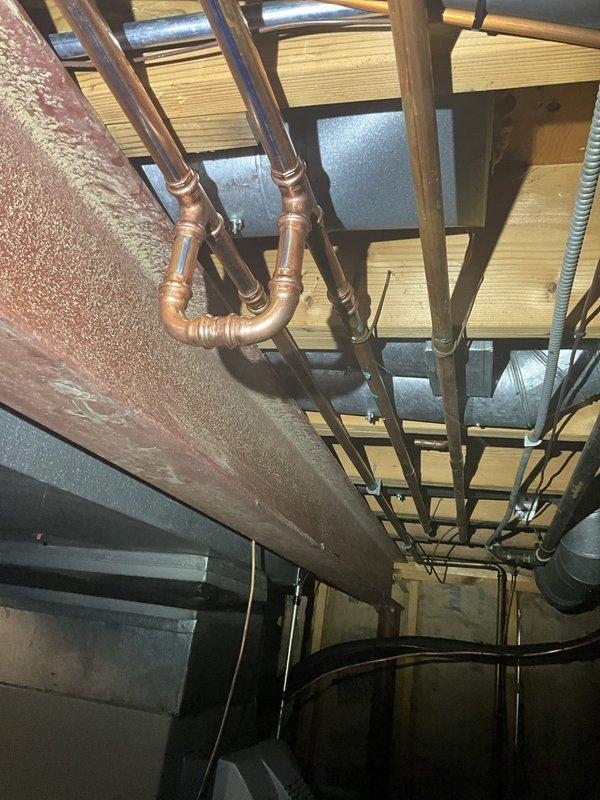 Responded to water leak call in basement near furnace following severe weather event. Upon inspection, identified pinhole leak in copper plumbing line running through floor joists in vicinity of 7-year-old heating unit. Photographically documented affected copper piping with U-bend configuration, soldered joints, and surrounding infrastructure including exposed joists and concrete block foundation walls. Noted moisture damage evident on adjacent wooden beam showing gray discoloration and deteriorated surface texture consistent with water exposure. Verified furnace operation normal with no impact to heating or cooling performance. Assessed plumbing system integrity and provided recommendations for copper line repair to prevent further water damage to structural components and mechanical equipment.