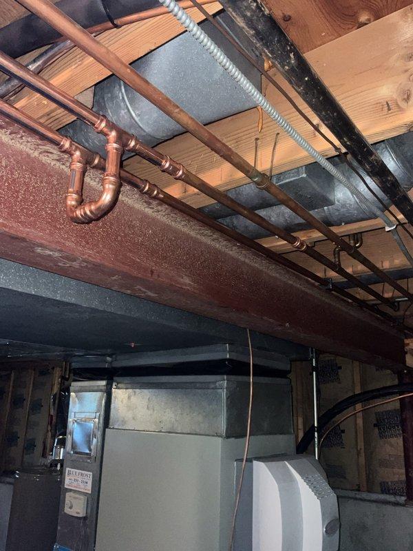 Responded to water leak call in basement near furnace following severe weather event. Upon inspection, identified pinhole leak in copper plumbing line running through floor joists in vicinity of 7-year-old heating unit. Photographically documented affected copper piping with U-bend configuration, soldered joints, and surrounding infrastructure including exposed joists and concrete block foundation walls. Noted moisture damage evident on adjacent wooden beam showing gray discoloration and deteriorated surface texture consistent with water exposure. Verified furnace operation normal with no impact to heating or cooling performance. Assessed plumbing system integrity and provided recommendations for copper line repair to prevent further water damage to structural components and mechanical equipment.