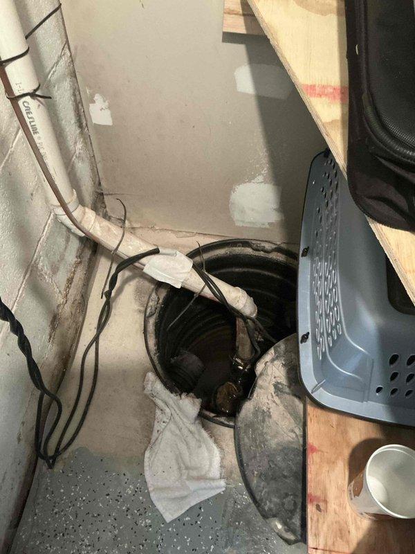 Completed comprehensive sump pump inspection in below-grade utility area. System evaluated included submersible pump unit with corrugated drainage inlet line and vertical discharge pipe showing rust staining on exterior surfaces. Pump housing, mounting hardware, and basin liner were inspected for proper operation and structural integrity. All components found functional with discharge piping secured by bracket assembly, though exterior corrosion noted on metal surfaces consistent with age and moisture exposure typical of below-grade water management installations.