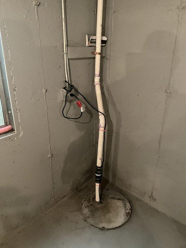Performed complimentary sump pump inspection in basement utility area. Examined PVC discharge piping assembly running from floor to ceiling, verified proper installation of pipe connections and supports, and assessed pump electrical connections. Inspected sump basin and pump unit, noting operational pump mechanism with standard metal housing and discharge piping configuration. Water level in pit was within normal operating range, and basin walls showed typical aging and mineral deposits consistent with regular use. System components appeared functional with no immediate concerns identified during visual inspection.