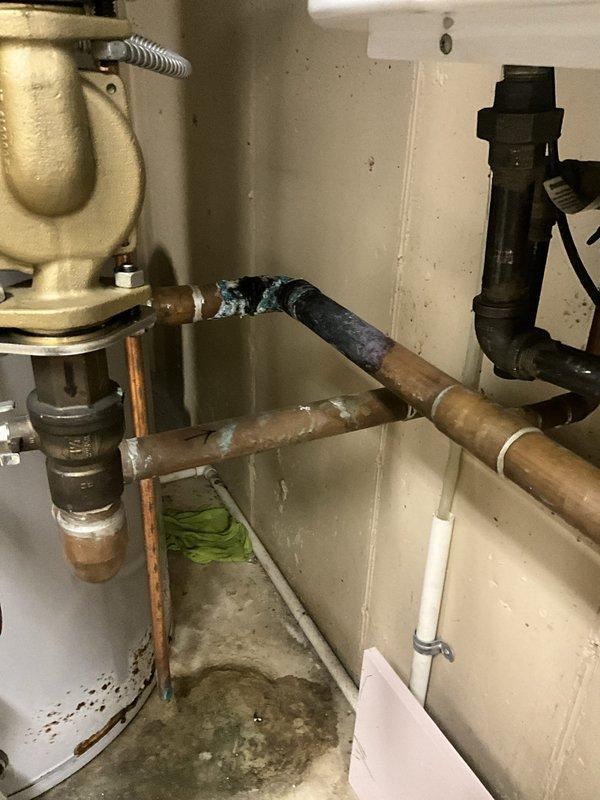 Responded to no hot water complaint at commercial facility. Upon inspection, found deteriorated copper piping with significant corrosion and joint failure near the A.O. Smith tanked water heater system. Evidence of active water leakage was present on concrete floor beneath compromised piping connections. Assessed water heater components, circulation pump, and associated plumbing infrastructure. Documented existing conditions showing darkened deterioration at pipe joints and moisture damage indicating ongoing leak. System evaluated for proper operation and identified failed piping as primary cause of service interruption.