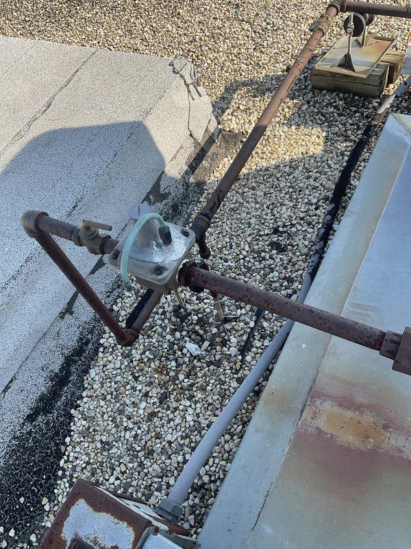 Conducted site assessment for commercial rooftop unit replacement project on flat gravel roof. Documented existing RTU installation including metal framework, ductwork assemblies, and associated piping systems showing extensive weathering and corrosion. Photographed rusted mounting brackets, oxidized pipe connections with teal-colored valve components, and deteriorated structural supports to determine replacement specifications. Evaluated rooftop access conditions and operational constraints for equipment installation during designated business hours from early morning through evening operations.