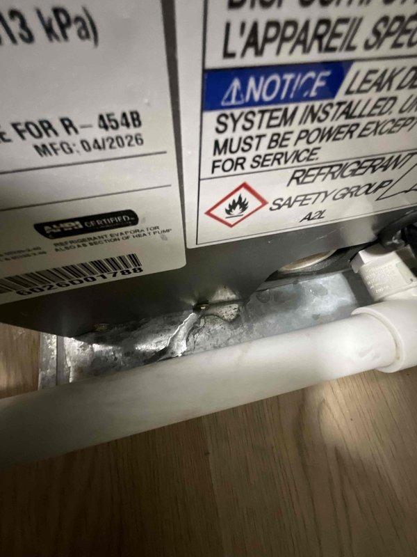 Responded to service call for water pooling under four-month-old furnace near AC drain line. Upon inspection, identified constant leak originating from condensate drainage system. Examined HVAC equipment including R-454B refrigerant system with A2L safety classification manufactured in April 2026. Inspected internal components including evaporator coil assembly with copper refrigerant lines, condensate pan, and drainage connections. Traced leak source to condensate drain line issue at PVC connection point. Documented equipment condition and drainage system configuration with photographs showing piping connections, coil assembly, and affected area. Cleared blockage and verified proper drainage flow through white PVC condensate line to eliminate standing water beneath unit.