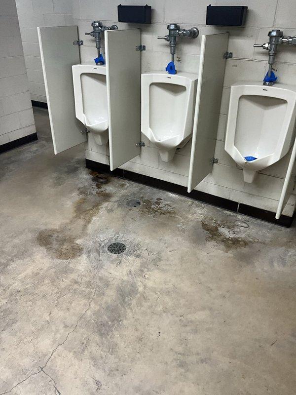 Responded to report of water intrusion through plumbing pipes in commercial restroom facility. Upon arrival, found evidence of water accumulation on concrete floor near urinal fixtures and floor drain, with visible staining and moisture patterns indicating recent or ongoing leak. Inspected three wall-mounted urinals with chrome flush valves and associated supply piping for source of water intrusion. Bathroom stalls cordoned off with caution tape due to facility conditions. Documented existing floor discoloration, water stains, and drain location for assessment. Investigation completed and photographic documentation obtained for further evaluation of plumbing system issues.