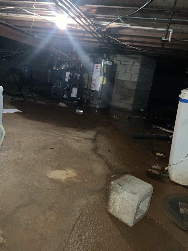 Conducted comprehensive assessment of basement mechanical systems including existing boiler and water heater installation. Documented significant moisture intrusion on concrete floor with visible water pooling and staining throughout the space, indicating potential drainage or groundwater issues that may impact equipment longevity. Evaluated dark blue boiler unit and gray cylindrical water heater, both with copper piping connections running to overhead plumbing along exposed ceiling joists. Photographed current equipment configuration, wet floor conditions, and overall installation environment to provide accurate proposal for system replacement and potential upgrades to address moisture concerns and improve heating efficiency.