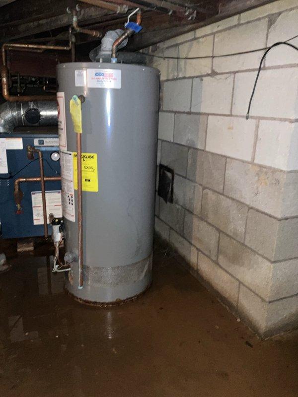 Conducted comprehensive assessment of basement mechanical systems including existing boiler and water heater installation. Documented significant moisture intrusion on concrete floor with visible water pooling and staining throughout the space, indicating potential drainage or groundwater issues that may impact equipment longevity. Evaluated dark blue boiler unit and gray cylindrical water heater, both with copper piping connections running to overhead plumbing along exposed ceiling joists. Photographed current equipment configuration, wet floor conditions, and overall installation environment to provide accurate proposal for system replacement and potential upgrades to address moisture concerns and improve heating efficiency.
