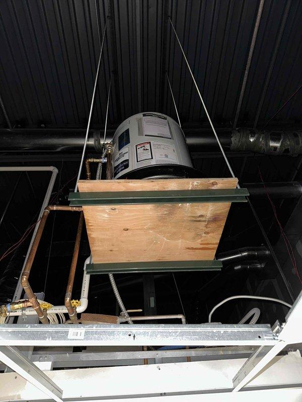 Installed new 20-gallon Bradford White electric water heater in ceiling location, replacing previous leaking unit. Mounted water heater on reinforced plywood platform with green metal channel supports, suspended by metal cables from the ceiling structure. Added boiler drain in ceiling for proper drainage and completed all necessary copper pipe connections with appropriate fittings.