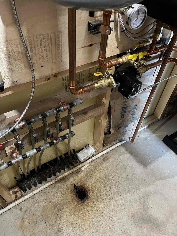 Responded to report of leaking black substance from accessory tank connected to IBC boiler system. Upon inspection, identified multiple leak sources including expansion tank with corrosion penetration above diaphragm releasing antifreeze sludge, aged indirect water heater with internal leak, and corroded connections at infloor heating manifold. Performed immediate repair on expansion tank to stop active leakage. System includes teal circulator pump, brass and copper piping with PEX connections, pressure gauge, and multi-zone manifold distribution assembly for radiant floor heating. Customer advised of additional repairs needed for water heater replacement and manifold connection rehabilitation, estimates to follow.