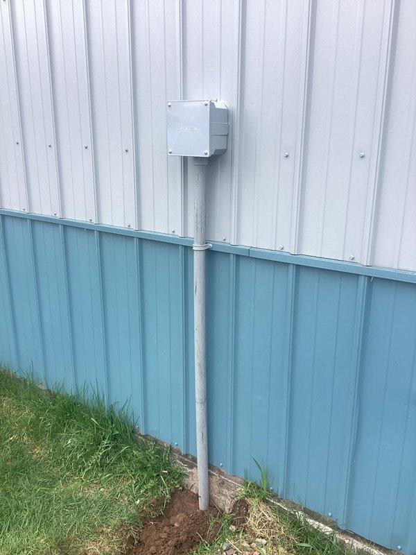 Removed existing 50A welder receptacle from building exterior and installed junction box to extend the circuit. Mounted 30/20 RV service panel on treated wooden post in yard area, running conduit from the building-mounted junction box down through the corrugated metal siding to ground level and across to the post-mounted panel. Connected the extended circuit to the RV panel and installed the required breaker to complete the electrical service, as the circuit was previously disconnected.