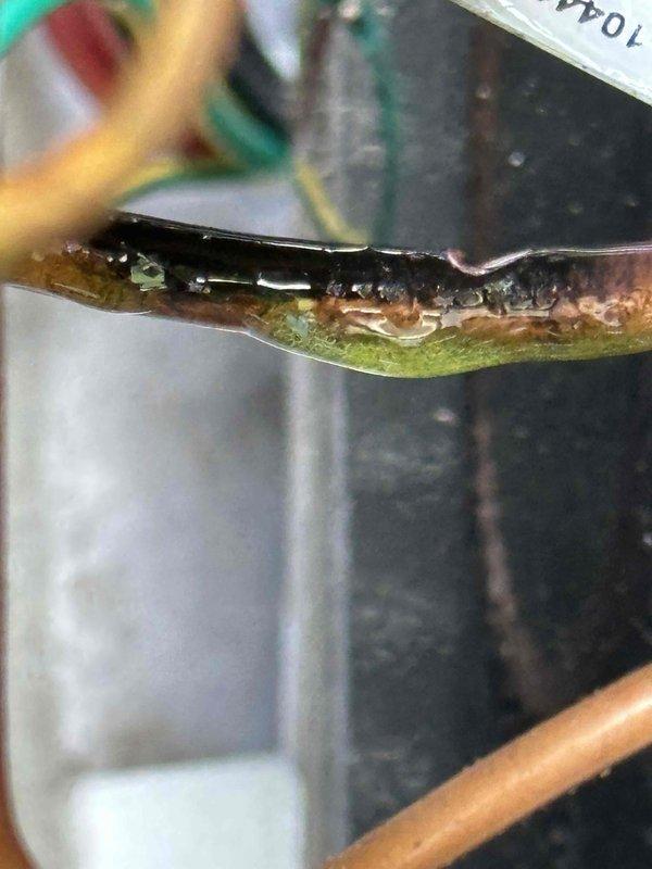 # Service Report: HVAC No Cooling Repair

Responded to service call for 9-year-old residential air conditioning system with no cooling condition and blinking red diagnostic light on outdoor unit. Upon arrival, performed visual inspection and diagnostic assessment of the condensing unit.

Removed outdoor unit access panel to inspect internal components and identify source of system failure. Located compressor assembly, refrigerant lines, condenser coil, and electrical connections. Observed blinking red diagnostic code indicating system fault condition.

During detailed inspection of refrigerant circuit, discovered significant deterioration and failure of copper suction line. Identified longitudinal split in copper tubing with visible corrosion, green patina from copper oxidation, and compromised pipe integrity. The damaged section exhibited brown discoloration and dark patches indicating advanced corrosion. This refrigerant line failure resulted in complete loss of refrigerant charge an