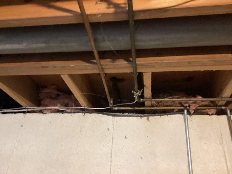 Completed comprehensive electrical maintenance home audit of unfinished utility space, inspecting exposed ceiling infrastructure including wooden joists, beams, and structural framing. Evaluated existing electrical systems including CFL lighting fixture with black circular mount, white electrical conduit routing and fastening methods along ceiling joists, and various electrical wiring running through framing members. Assessed HVAC ductwork positioning and metal support straps, verified proper clearances between electrical components and mechanical systems including gray rectangular ductwork and copper piping. Documented exposed infrastructure conditions, insulation placement between framing members, and overall electrical installation compliance with building codes. Photographed all findings for maintenance records.