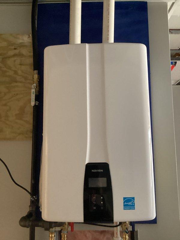 Completed full installation of commercial-grade Navien tankless water heater system with proper venting and plumbing connections. Mounted unit on plywood backing against blue foam insulation board, installed three white PVC vent pipes for exhaust and air intake, and connected copper supply lines with brass fittings and shut-off valves for hot and cold water service. Established electrical connection and verified digital control panel functionality upon completion.