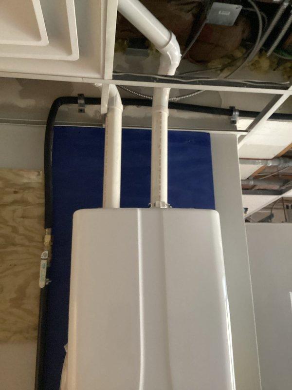 Completed full installation of commercial-grade Navien tankless water heater system with proper venting and plumbing connections. Mounted unit on plywood backing against blue foam insulation board, installed three white PVC vent pipes for exhaust and air intake, and connected copper supply lines with brass fittings and shut-off valves for hot and cold water service. Established electrical connection and verified digital control panel functionality upon completion.