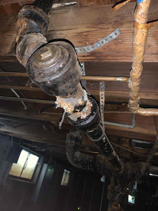 Responded to kitchen sink clog complaint and performed diagnostic inspection of drainage system. Upon investigation, accessed basement plumbing infrastructure and documented existing conditions of cast iron drain lines and associated piping components. Inspection revealed aging plumbing system with visible corrosion on cleanout fittings and drain assemblies beneath kitchen area. Assessed drainage configuration and confirmed presence of exterior sewer cleanout as reported. Photographed plumbing conditions including corroded cast iron piping, cleanout access points, and overall drainage layout for service records. Kitchen sink drainage issue addressed and system restored to proper working order.