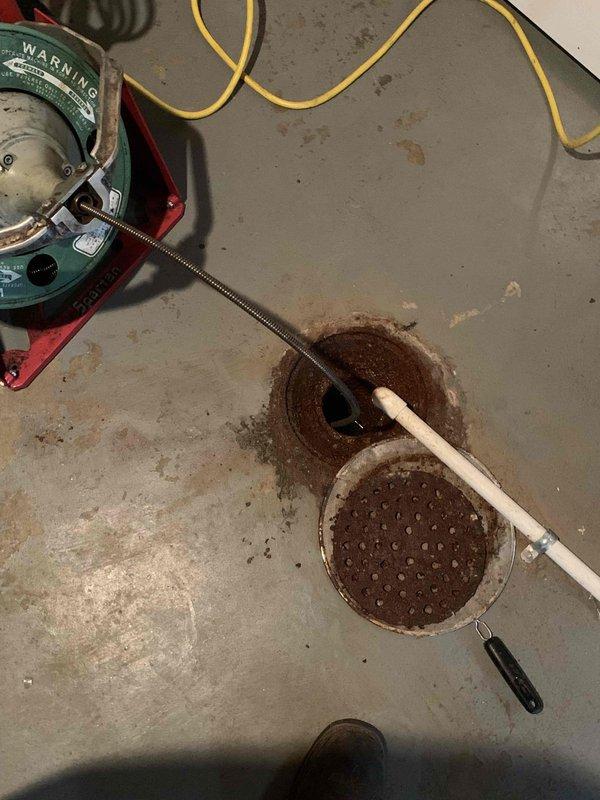 Responded to main sewer line backup in basement. Upon arrival, located floor drain with visible sewage backup and sediment accumulation. Removed drain cover revealing significant debris buildup coating interior drain walls and dark brown sediment obstruction. Deployed mechanical drain auger with flexible cable through floor drain opening to clear main line blockage. Successfully broke through obstruction and restored proper drainage flow. Removed accumulated debris and sediment from drain system. Tested drain flow to confirm clearance of main sewer line. Left drain operational with proper flow restored.
