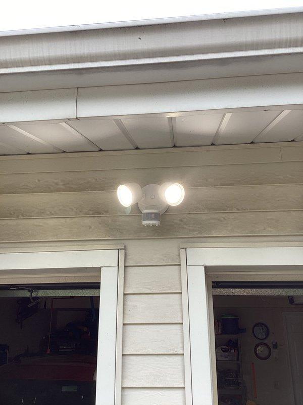 Completed installation of electrical lighting fixtures for residential property. Installed dual-head motion-sensor security light fixture on exterior wall near garage area with proper mounting and electrical connections. Added interior garage electrical components including light switch with cover plate and duplex outlet with cover plate mounted on wooden post near garage door frame. All fixtures tested and verified operational upon completion.