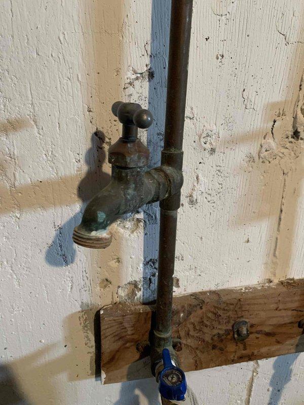 Responded to a plumbing service call for a clogged drain located downstairs near the washing machine that was backing up during laundry cycles. Additionally addressed an unrelated water supply line leak that exhibited a steady drip. Inspected and photographed the exterior plumbing connections, including an aged outdoor spigot assembly showing significant corrosion and weathering, with visible oxidation on brass fittings and a blue-handled valve at the base connection point.