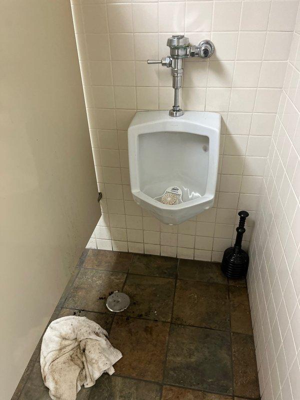 Responded to clogged drain issue in men's restroom facility. Upon arrival, assessed both toilet and urinal fixtures for drainage problems. Utilized plunger to clear obstruction in floor drain system. Verified proper water flow and drainage function in all fixtures before completing service. Restroom returned to full operational status.